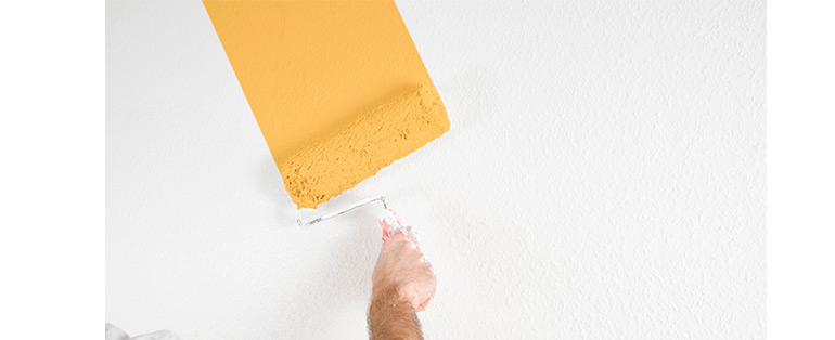 Imagem de mão caucasiana passando rolo de tinta na cor amarela em parede branca.
