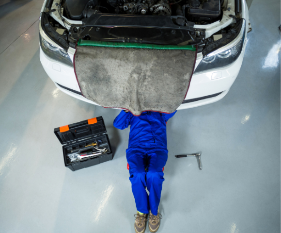 Técnico mecânico de uniforme azul em oficina, com carro com o capô aberto e ferramenta de trabalho ao lado, utilizando uma Esteira para mecânico para manutenção e inspeção do motor.